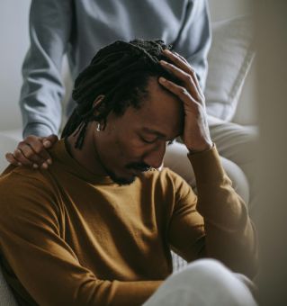 Worried depressed African American male touching head nervously and sitting with eyes closed near supporting wife in bedroom