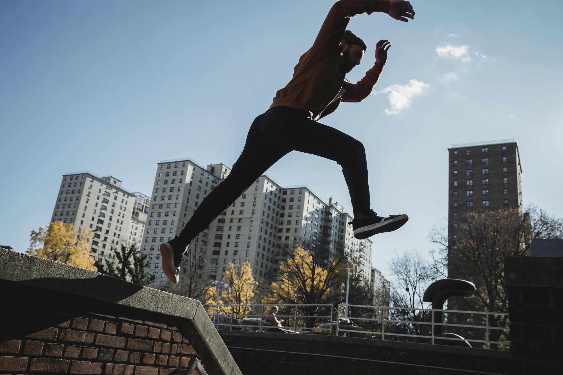 Side view energetic young male tracer in casual clothes jumping from brick fence against modern city buildings on clear sunny day