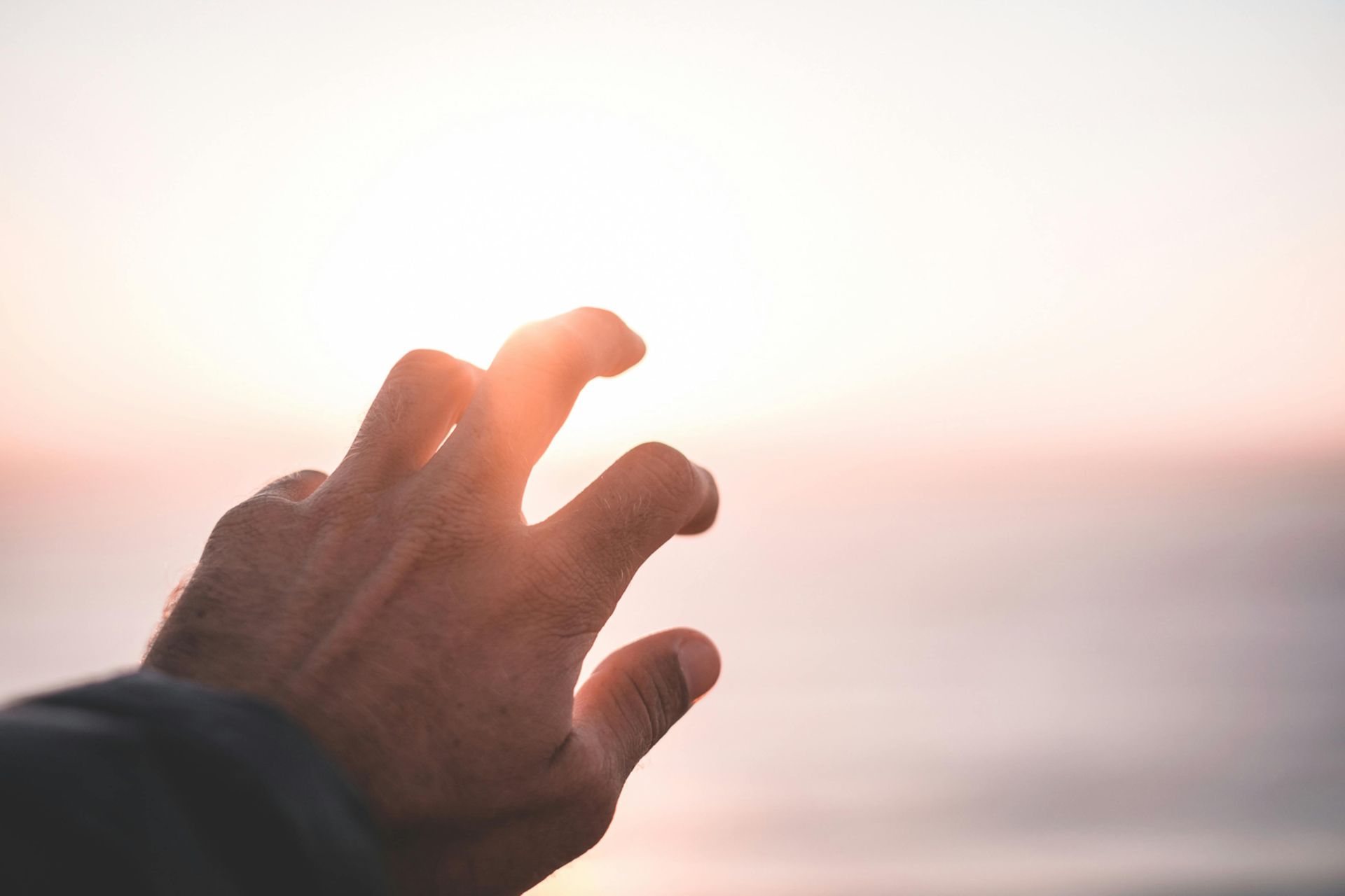 Crop anonymous male reaching hand to sun while standing near sea at sundown